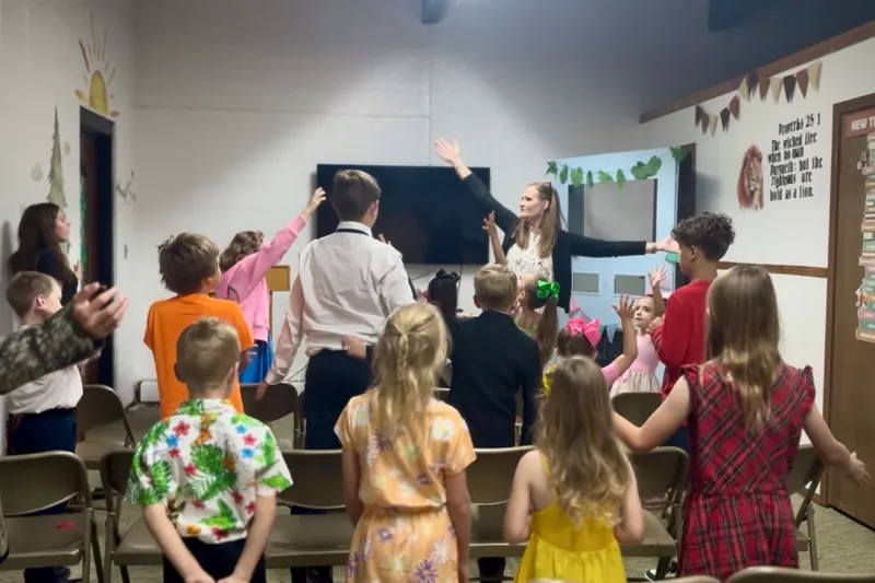 Children standing with their hands raised in song while a teacher leads from the front of the Jr Church room.
