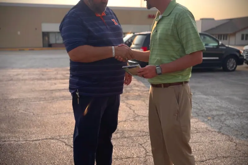 Two men shaking hands in a car park at sunset, one handing the other a gospel tract.