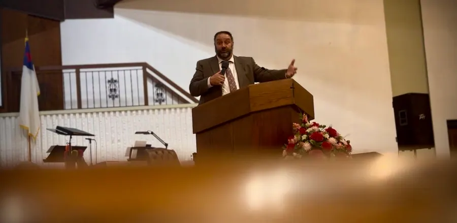 Pastor Paul Coyle preaching from the pulpit with a microphone, viewed from the pews.